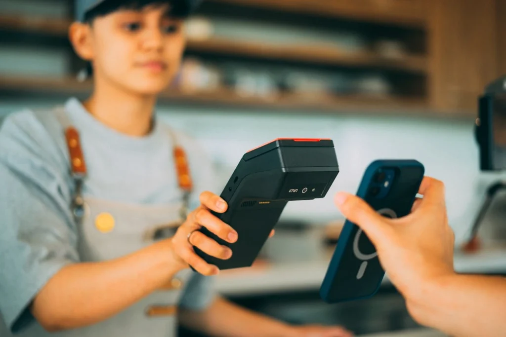 A customer using a smartphone for a contactless mobile payment on a handheld portable POS terminal held by a staff member.